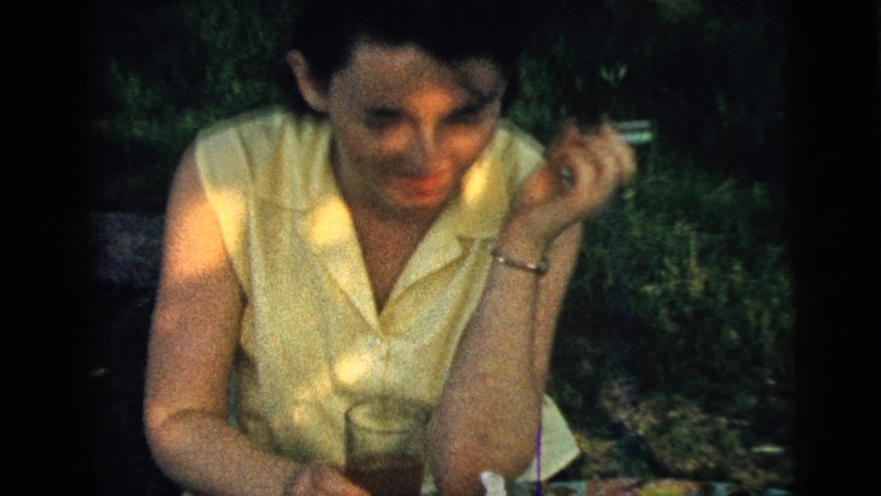 A woman in 1961 Camden home-movie footage smiles toward the camera while holding a cigarette beside a glass drink outdoors.