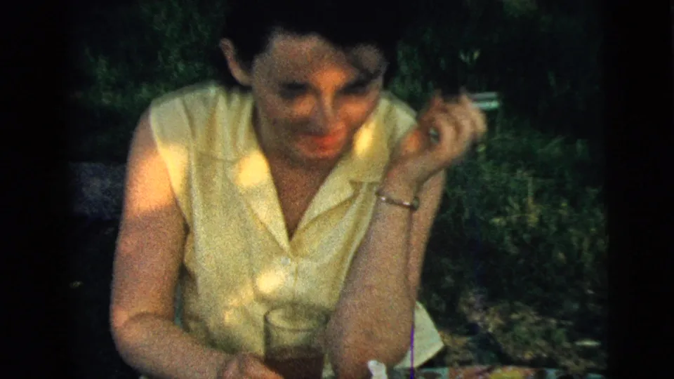 A woman in 1961 Camden home-movie footage smiles toward the camera while holding a cigarette beside a glass drink outdoors.
