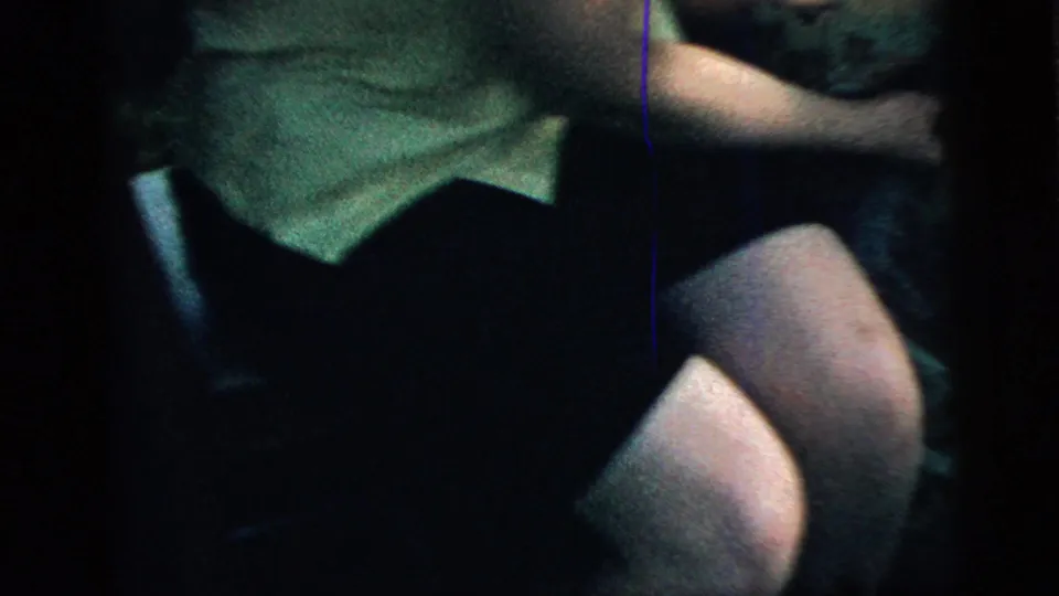 A close, low-framed home-movie view shows the seated woman in a yellow blouse, her legs, a chair, and another person partly visible nearby.