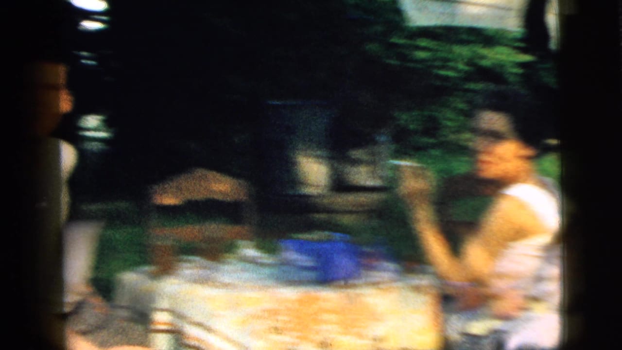 In the Camden 1961 backyard scene, adults sit around a table with a patterned cloth while a woman gestures with a cigarette near garden greenery.