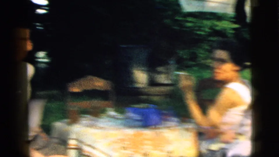 In the Camden 1961 backyard scene, adults sit around a table with a patterned cloth while a woman gestures with a cigarette near garden greenery.