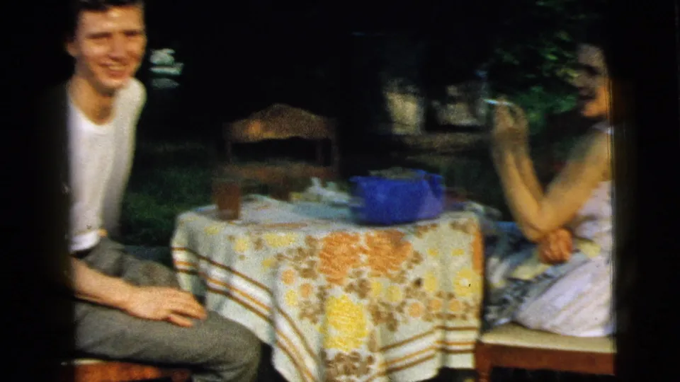 A young man in a white T-shirt sits beside a woman at a backyard table in Camden, New Jersey, with glasses, a blue container, and patterned cloth visible.