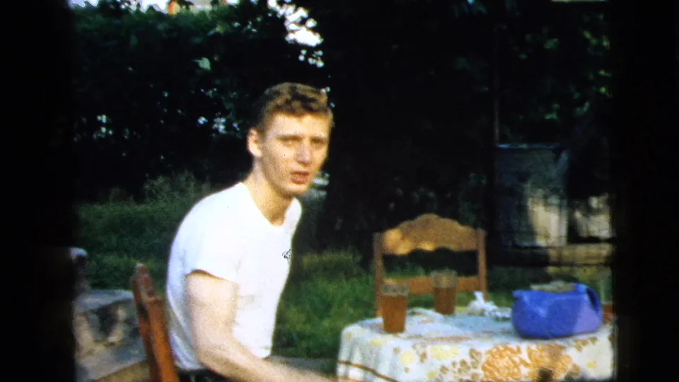A young man in a white T-shirt sits near a patterned backyard table in the 1961 Camden footage, with greenery, chairs, glasses, and a blue container nearby.