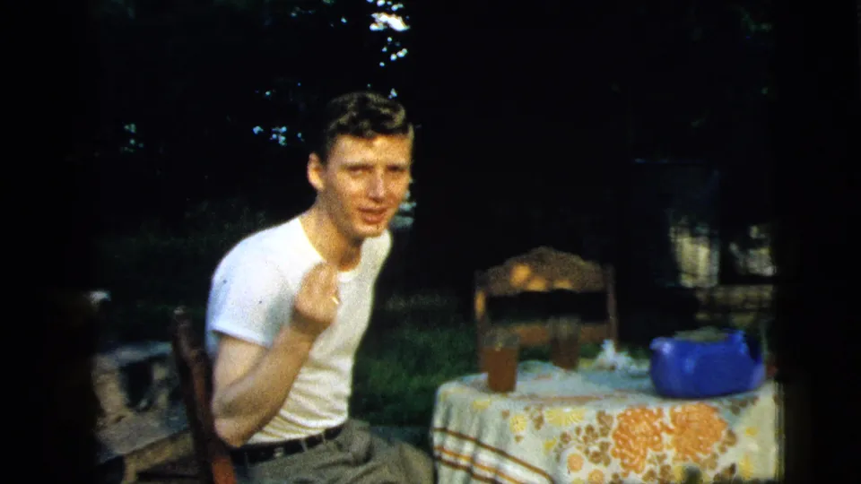 In 1961 Camden, New Jersey home-movie footage, a young man sits at an outdoor table with a floral cloth and blue teapot.