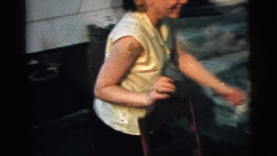 The young woman moves quickly past the dark car in the Camden 8mm footage, with house siding and a stone wall visible behind her.