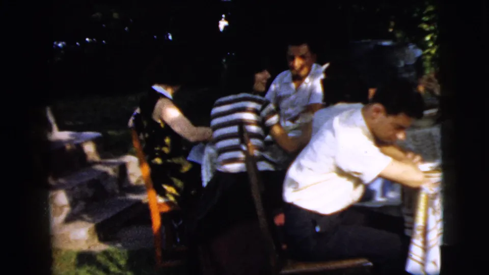 A Camden backyard gathering shows adults seated around a table, with chairs, dishes, greenery, and shaded 8mm color exposure.
