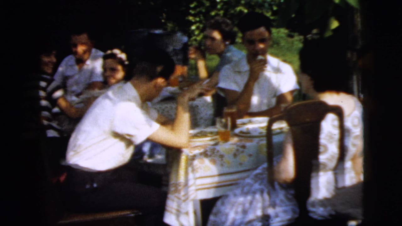 Several adults sit at a table outdoors, with a man leaning forward in the foreground.