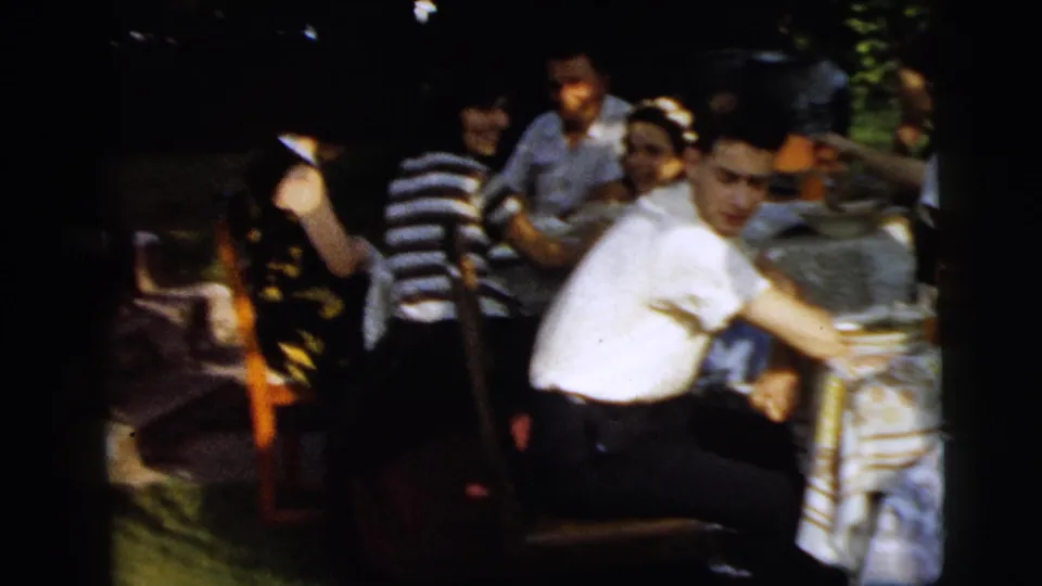Backyard meal scene in 1961 Camden with adults seated around a table, framed through dark film edges and foliage.