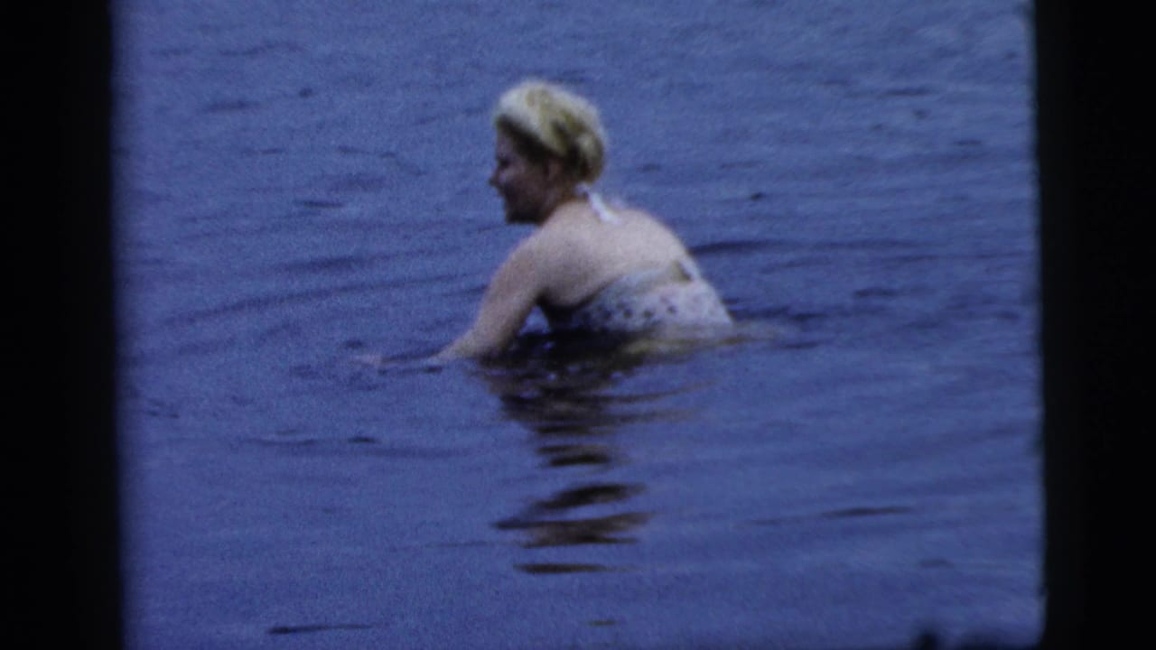 Adult blonde woman in a patterned bathing suit swims in blue-tinted water in 1961 Camden home-movie footage.