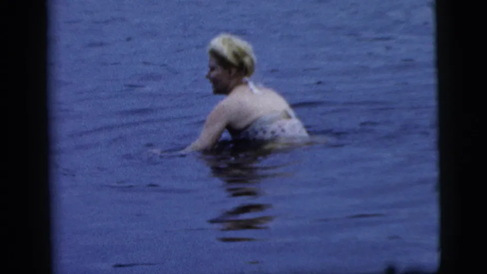 Adult blonde woman in a patterned bathing suit swims in blue-tinted water in 1961 Camden home-movie footage.