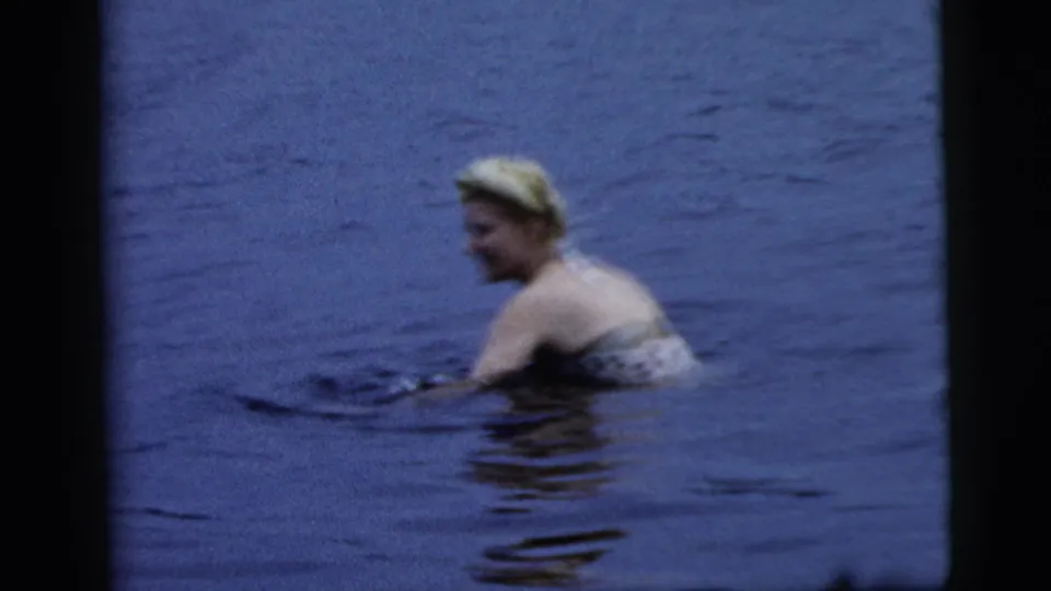 Adult blonde woman in a patterned bathing suit turns toward the camera while swimming in Camden footage from 1961.