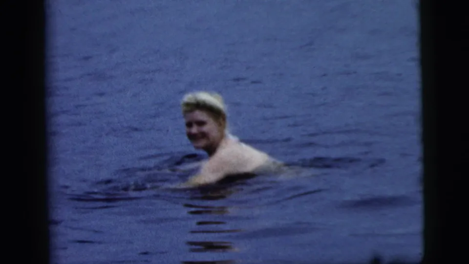 The woman continues swimming through blue lake water, glancing back toward the camera in the 1961 Camden home-movie footage.