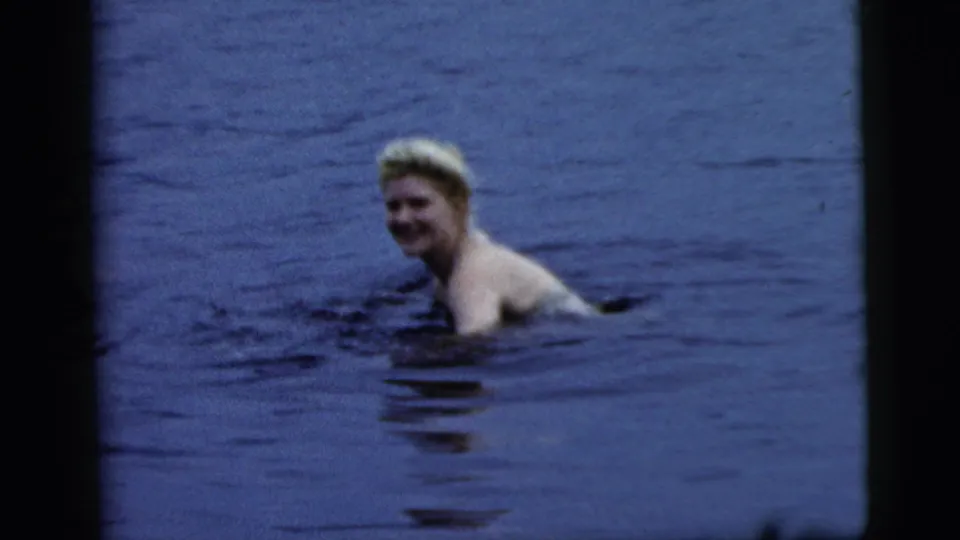 A smiling woman is partly submerged in lake water, framed softly by the 8mm scan and dark film edge.