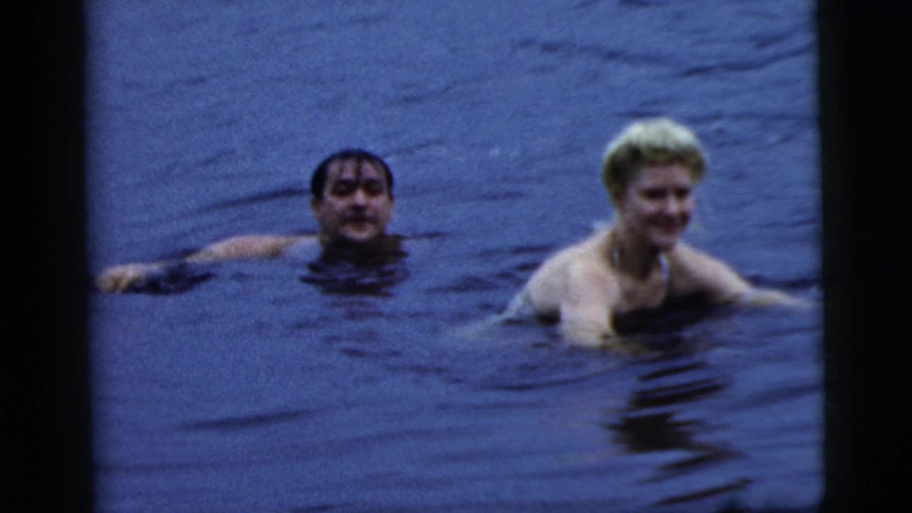 1961 Camden home-movie frame showing a brunet man and a blonde woman in a bathing suit swimming in dark water.