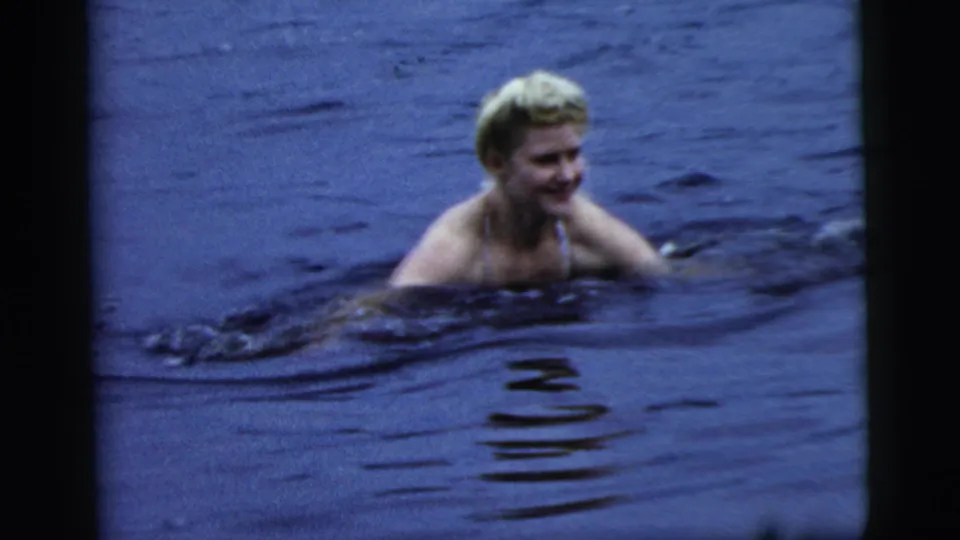 1961 Camden home-movie frame showing a blonde woman in a bathing suit swimming sideways through dark rippling water.