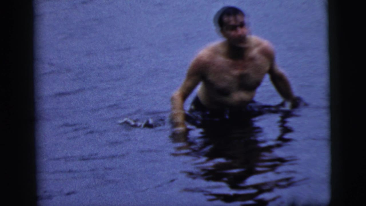 Shirtless man stands or wades chest-deep in lake water.