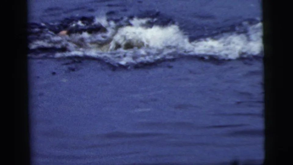 A swimmer moves low through the water, leaving a broad splash and wake in the vintage 8mm frame.