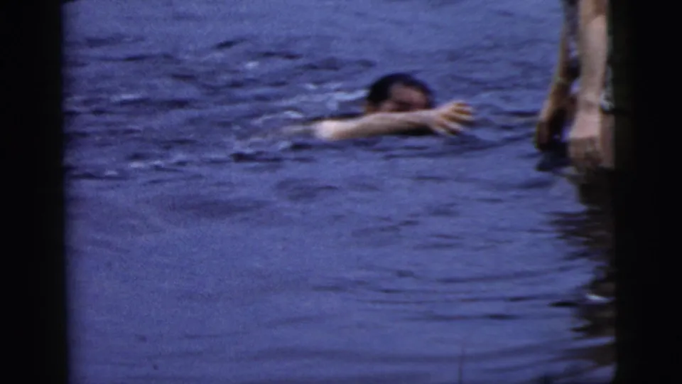 A swimmer reaches across the water toward a partially visible standing person in 1961 Camden home-movie footage.