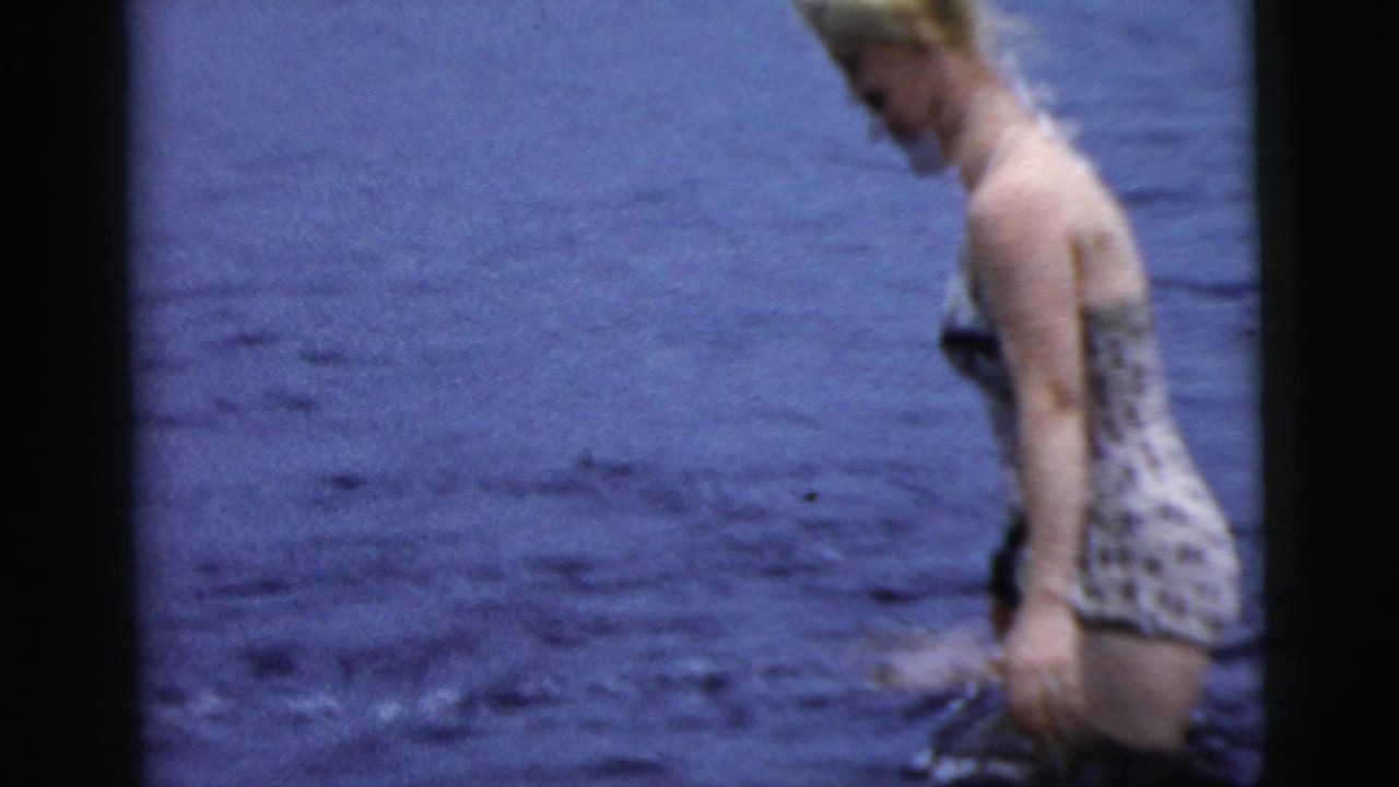 Blonde woman in a patterned swimsuit wading through dark lake water.