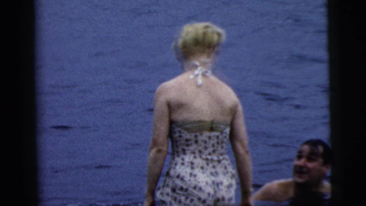 A woman in a patterned swimsuit stands in lake water while a man swims nearby in this Camden, New Jersey home-movie frame.