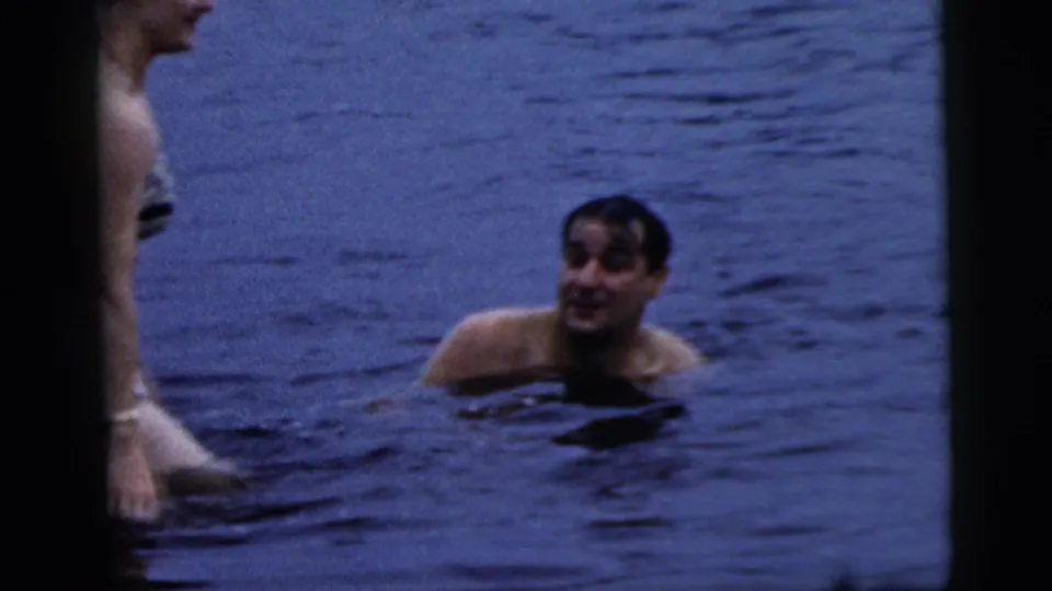 A man smiles while partly submerged in lake water, with the woman cropped at the left edge of the 1961 home-movie frame.
