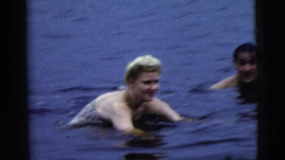 The blonde woman swims through the lake while a man appears at the right edge in the Camden, New Jersey 1961 footage.