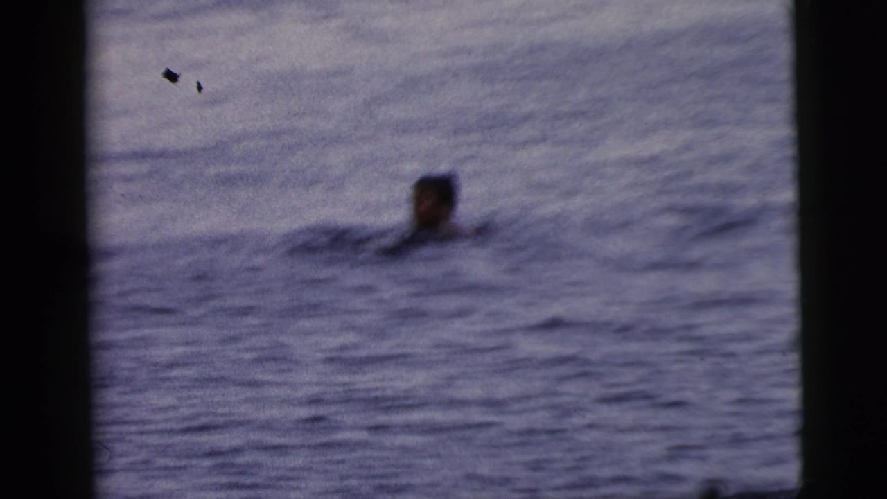 1961 Camden home-movie frame showing a distant swimmer's head above open water with dark film borders and soft focus.