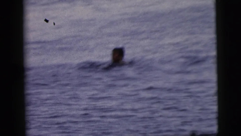 1961 Camden home-movie frame showing a distant swimmer's head above open water with dark film borders and soft focus.