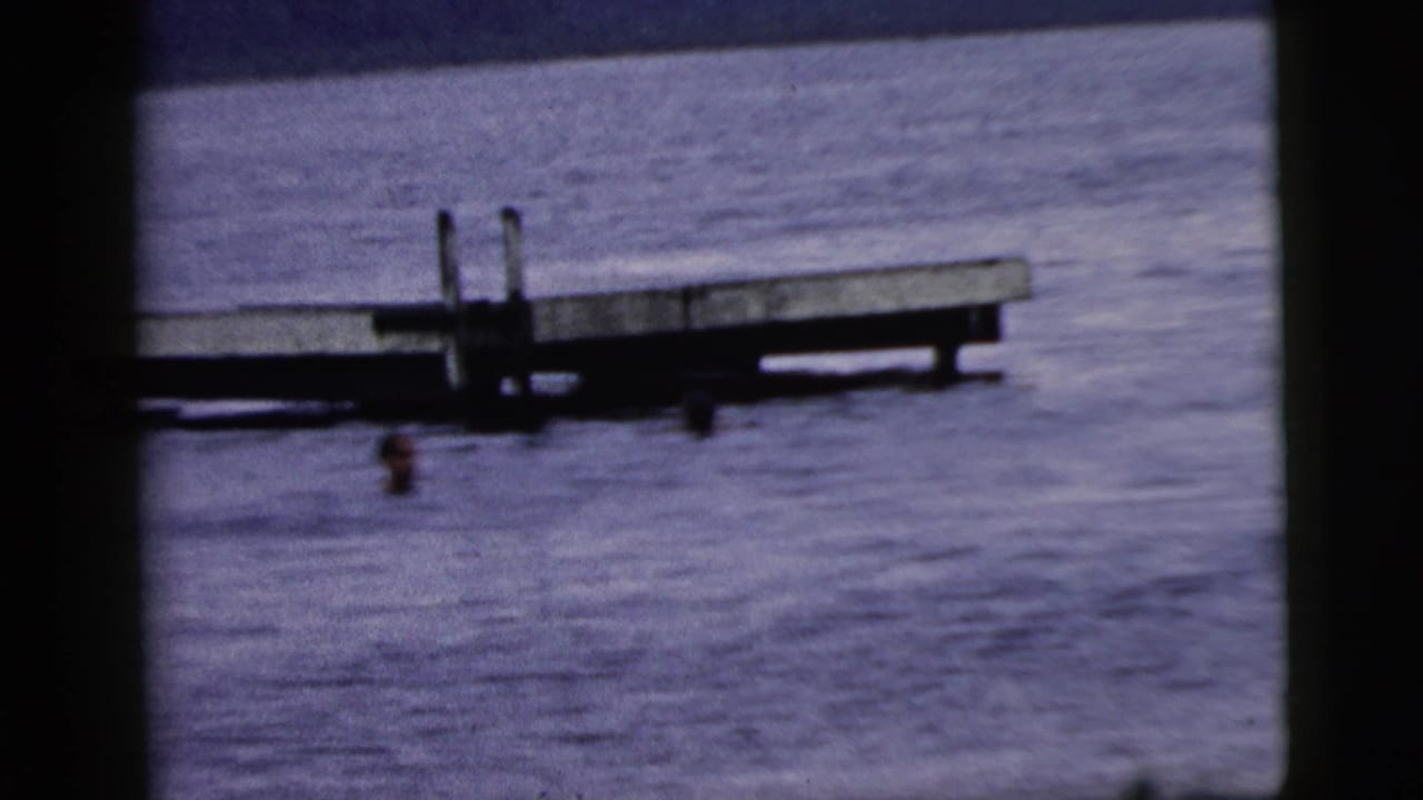 Home-movie frame from the 1961 Camden metadata set with swimmers near a wooden platform on open water.