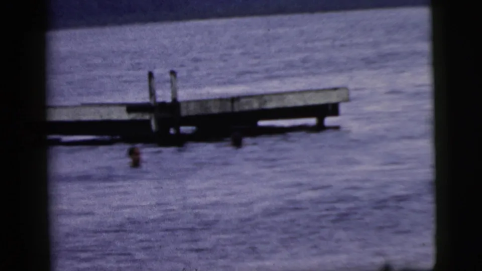 Home-movie frame from the 1961 Camden metadata set with swimmers near a wooden platform on open water.