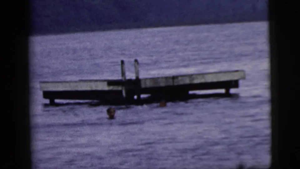 1961 8mm home-movie frame showing two swimmers beside a low wooden platform with a ladder in blue water.