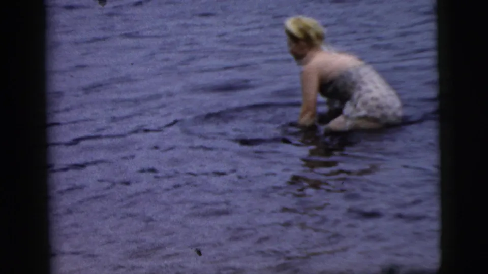 The woman appears to kneel or pause while wading in open water, seen through soft 1961 home-movie footage.