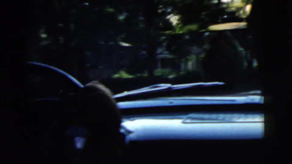 1961 Camden home-movie frame showing a dim car interior, steering wheel, dashboard, windshield, and leafy street beyond.