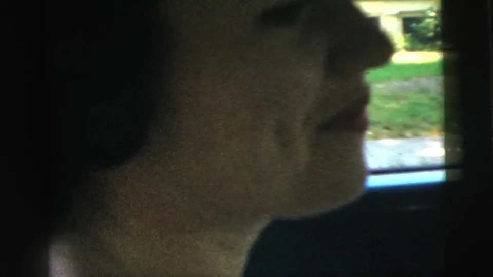 Woman in side profile inside a car in 1961 Camden, with soft daylight from the window and greenery outside.