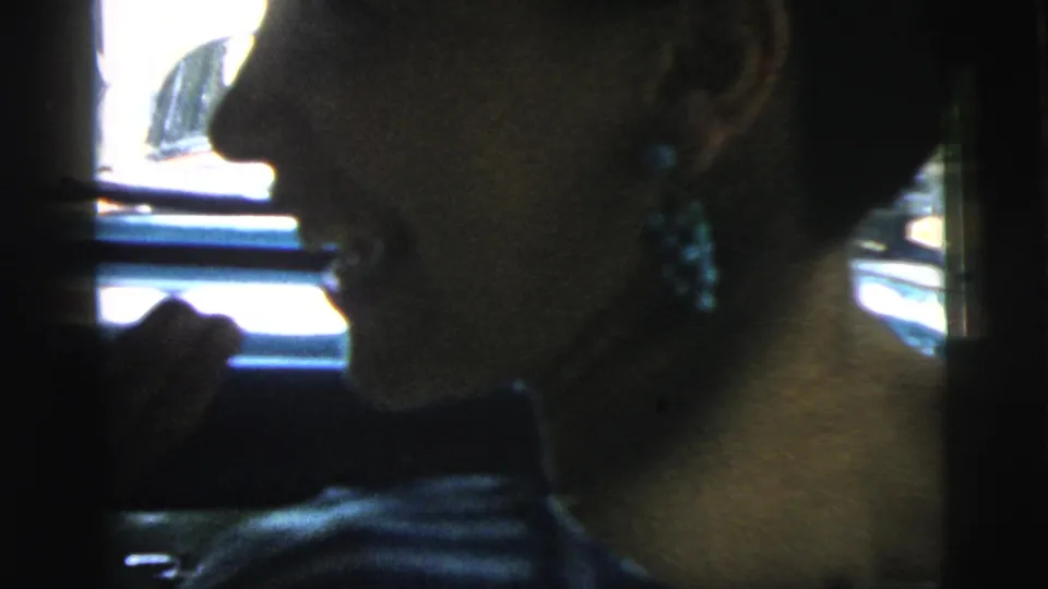 Woman in profile inside a car in 1961 Camden, appearing to smile or speak, with earrings and bright window light visible.