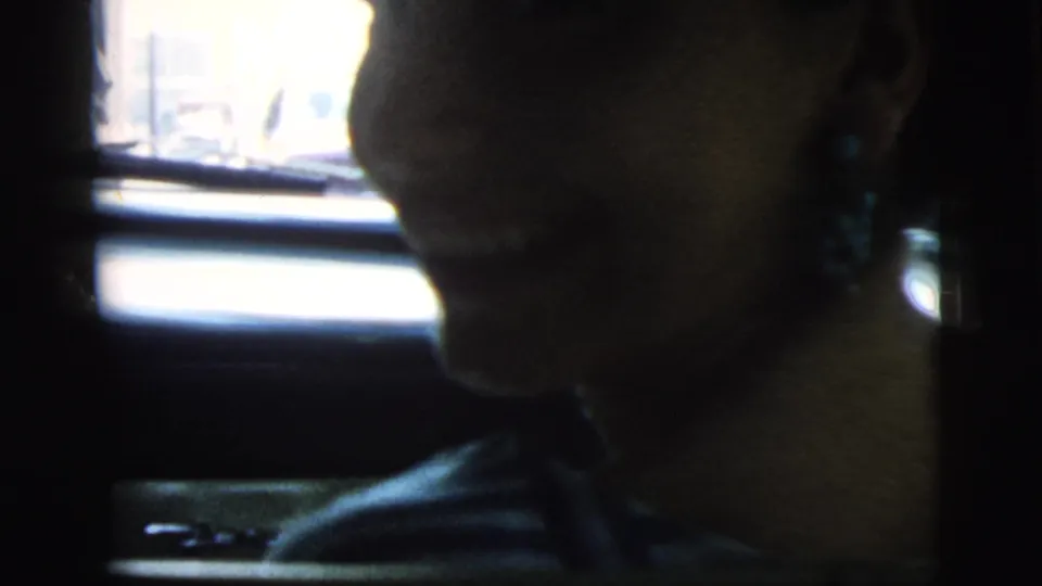The woman smiles faintly in profile inside the car, her face partly lit by daylight from the side window in the 1961 home movie.