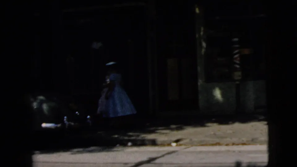 A woman in a full-skirted dress walks along a shaded sidewalk near storefront windows and a parked car.