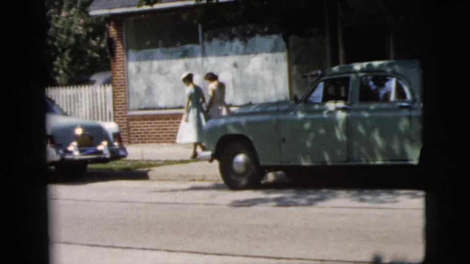 Two women in dresses and hats walk past a green vintage car on a Camden street in 1961.