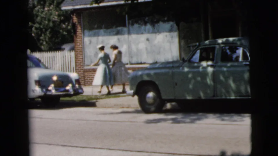 In Camden, New Jersey wedding footage from 1961, two women in full-skirted dresses walk past early 1960s cars on a neighborhood sidewalk.