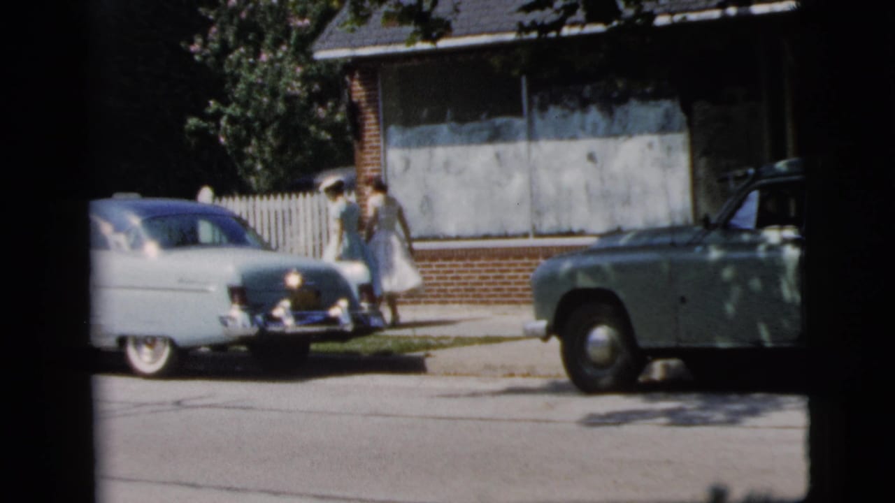 Two women stand near a white car while another car is parked at the curb.