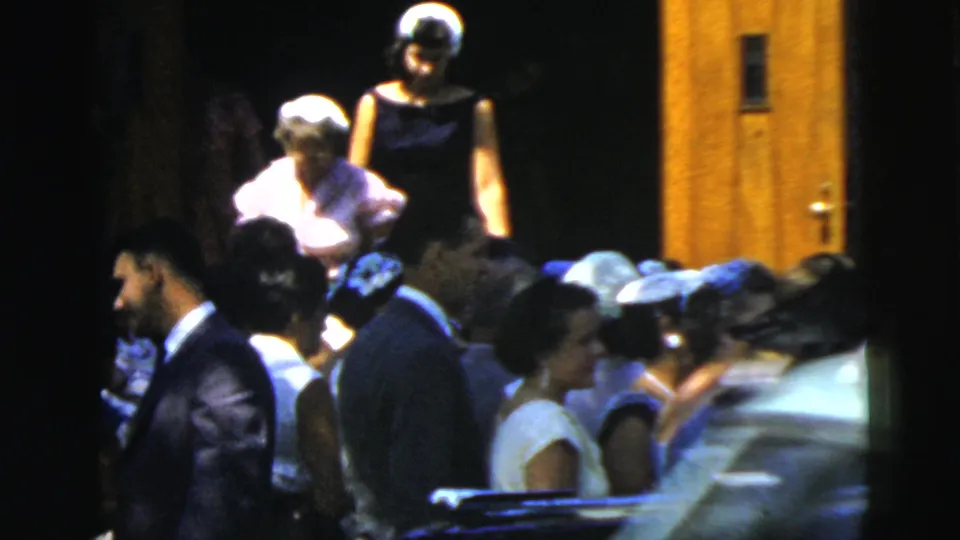 A crowded group of wedding guests in suits, dresses, and hats gathers near wooden doors in dim 8mm home-movie footage.