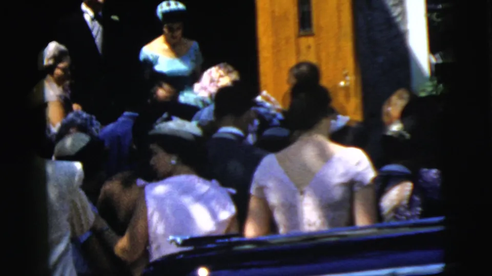 At the Camden wedding gathering, women in pastel dresses and hats stand in the foreground as guests fill the church entrance area.