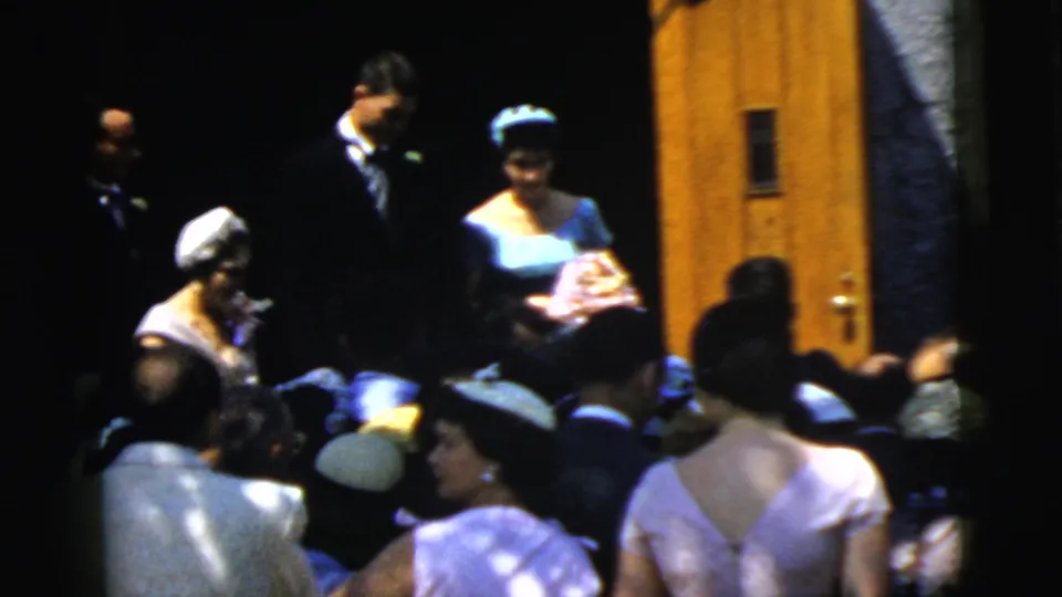 The bride and groom remain near the Camden church entrance in 1961 while formally dressed guests gather in the sunlit foreground.