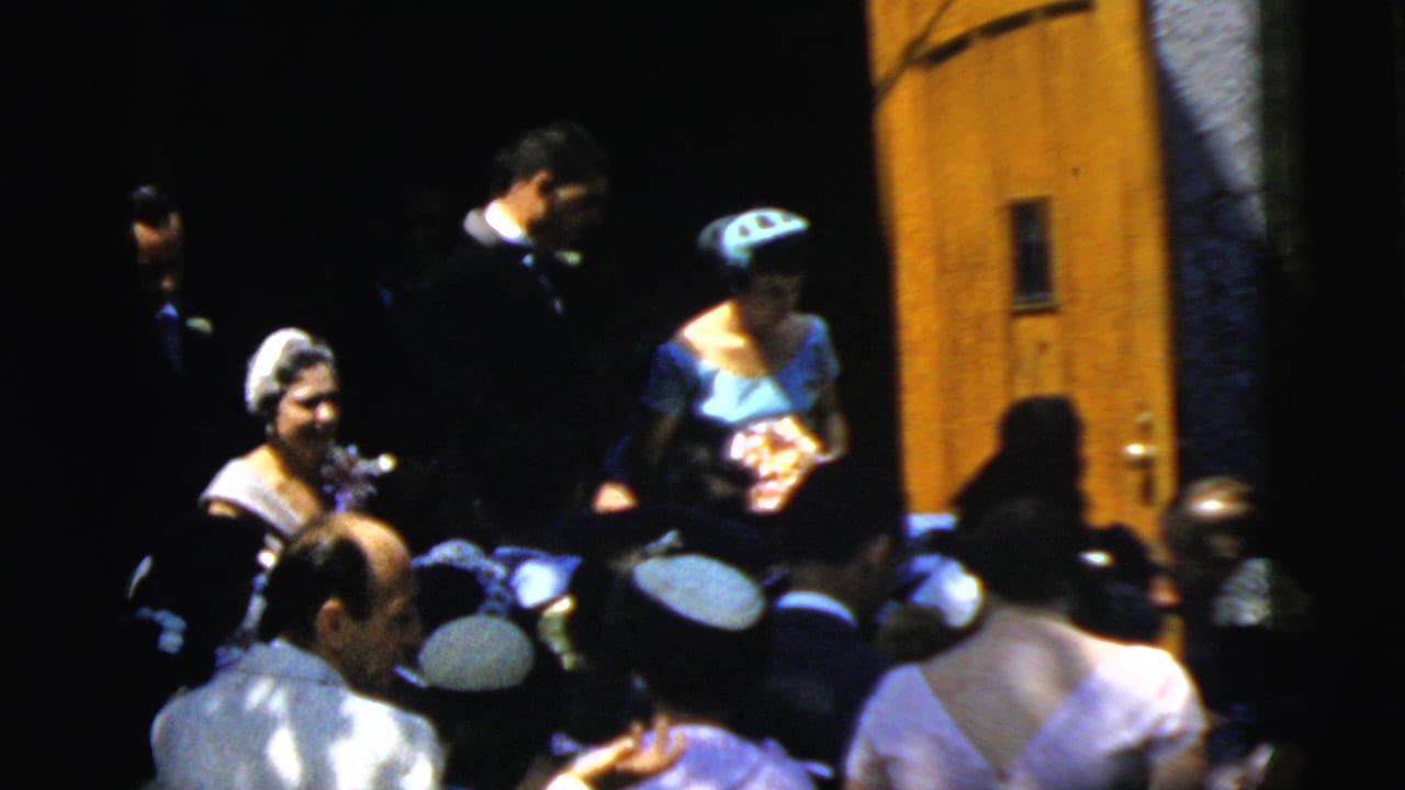 Wedding guests and attendants gather near a doorway in bright sunlight.