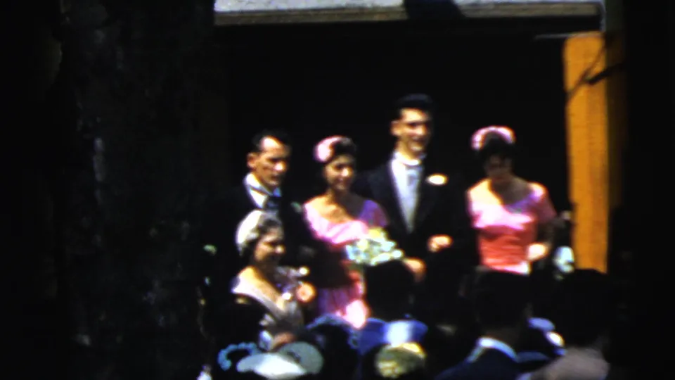 1961 Camden wedding home-movie frame showing the wedding party posed near a doorway with guests in the foreground.