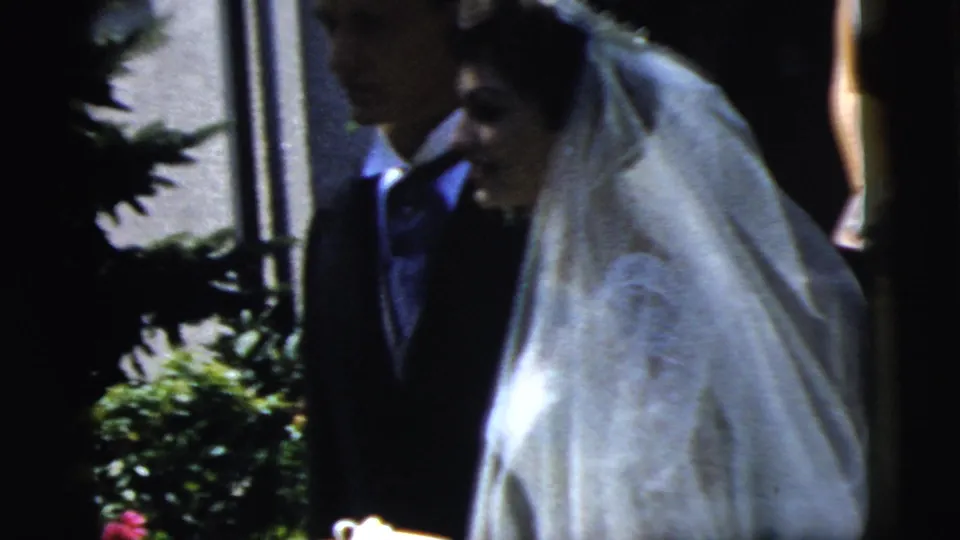 Bride in a white veil and groom in a dark suit move past a hedge outside the church during the Camden wedding scene.