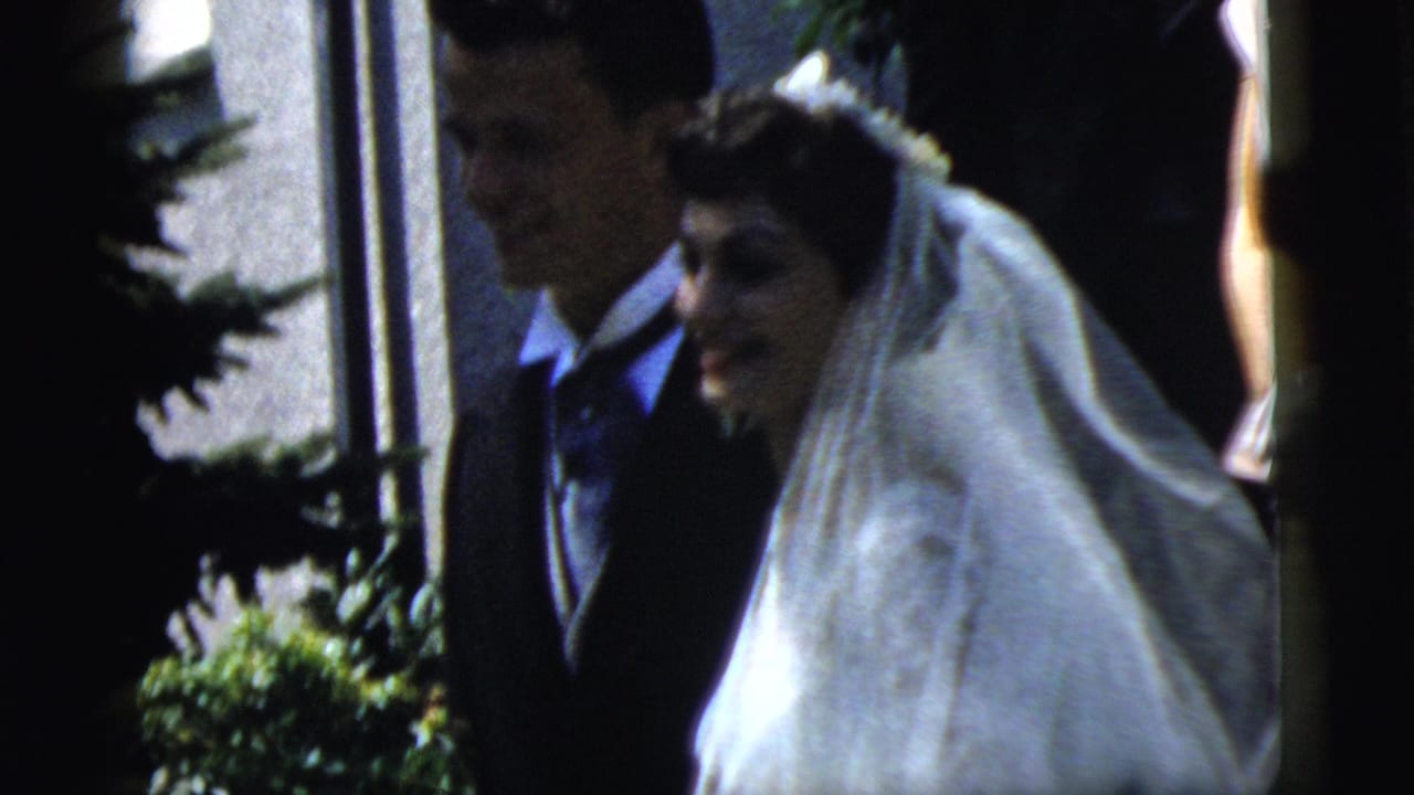 Bride in a white veil walks beside a groom in a dark suit outside a building.