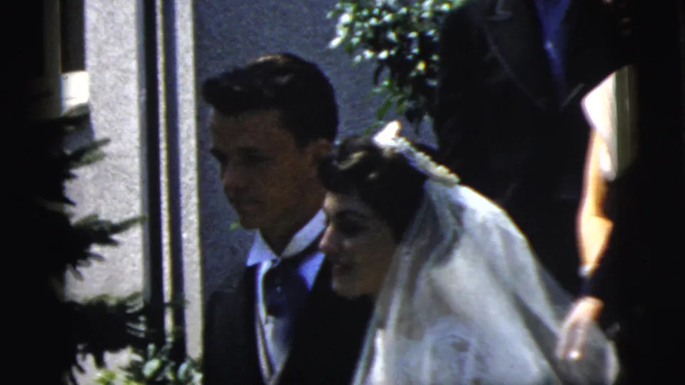 A groom in a dark suit and a bride in a white veil pass the camera outside a Camden building during the 1961 wedding footage.