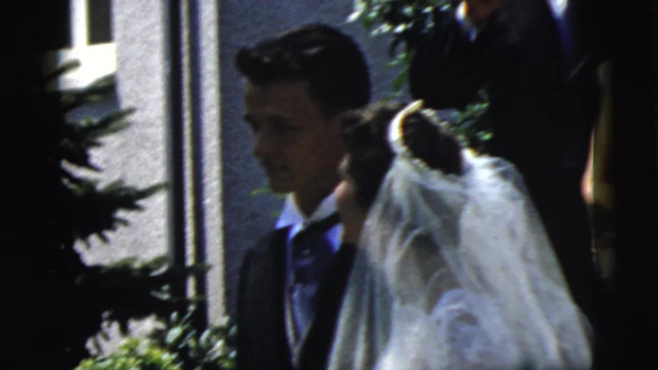 The groom looks left as the bride turns under her veil, with another adult partly visible near the building entrance in the 1961 Camden scene.