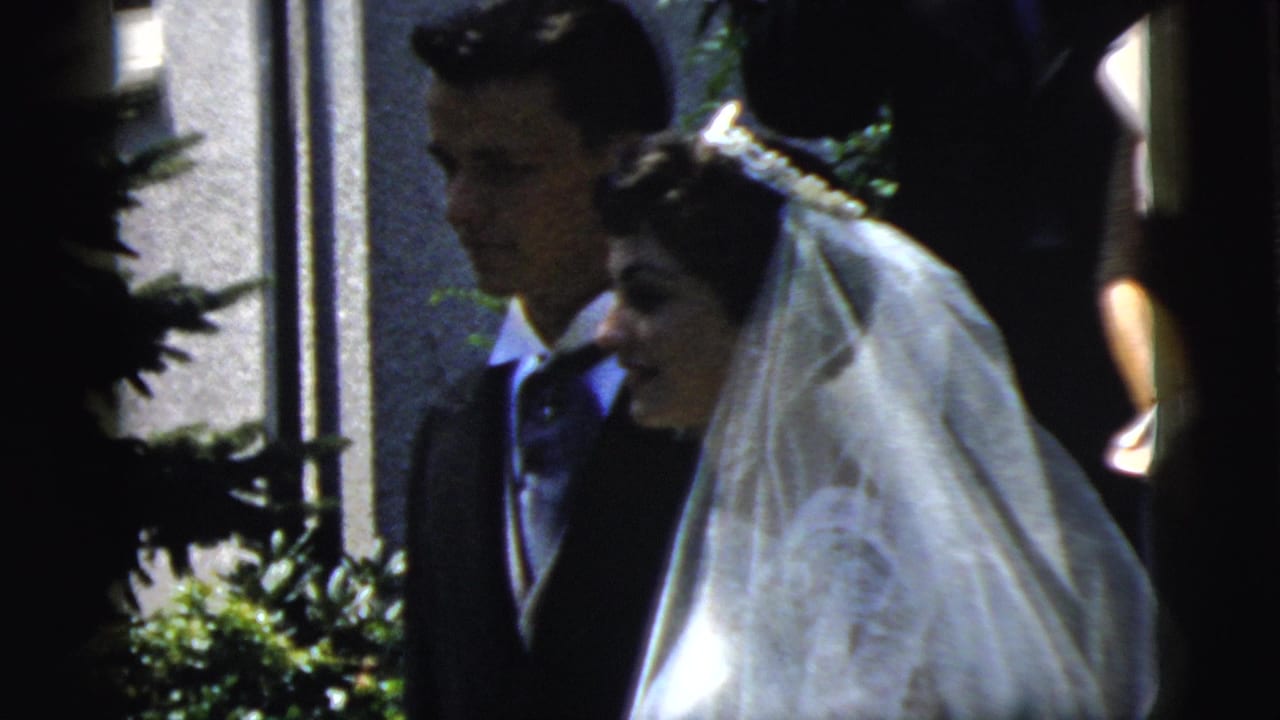 Bride in a white veil smiles beside a groom in dark formalwear near a doorway.